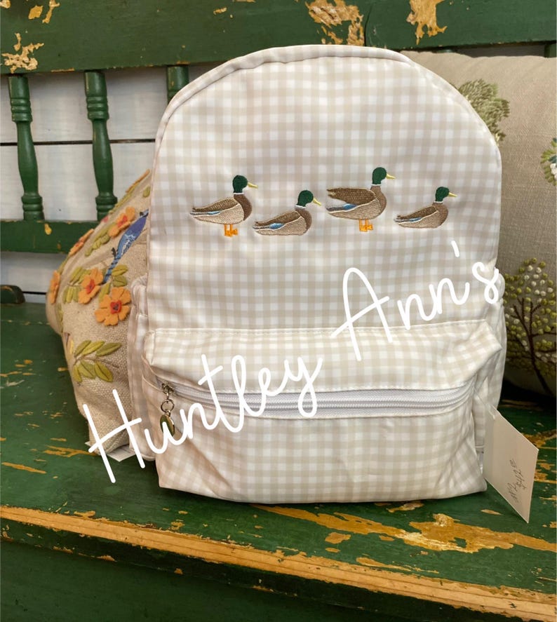 May include: A beige and white gingham backpack with embroidered ducks. The backpack has a zippered pocket and a small tag that says "Huntley Ann's".