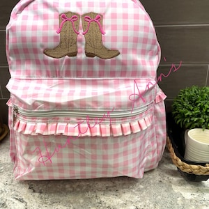 May include: A pink and white gingham patterned backpack with a zip pocket and frill trim. The design features two embroidered brown cowboy boots with pink bows. The top handle and zip are white.