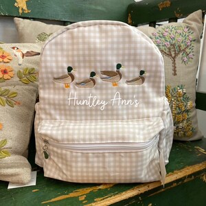 May include: A beige and white gingham backpack with a zippered pocket and a silver zipper pull. The backpack has an embroidered design of four ducks in a row and the text "Huntley Ann's".