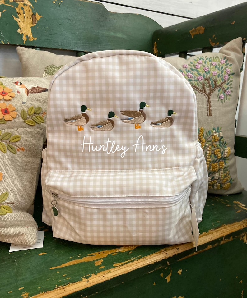 May include: A beige and white gingham backpack with a zippered pocket and a silver zipper pull. The backpack has an embroidered design of four ducks in a row and the text "Huntley Ann's".