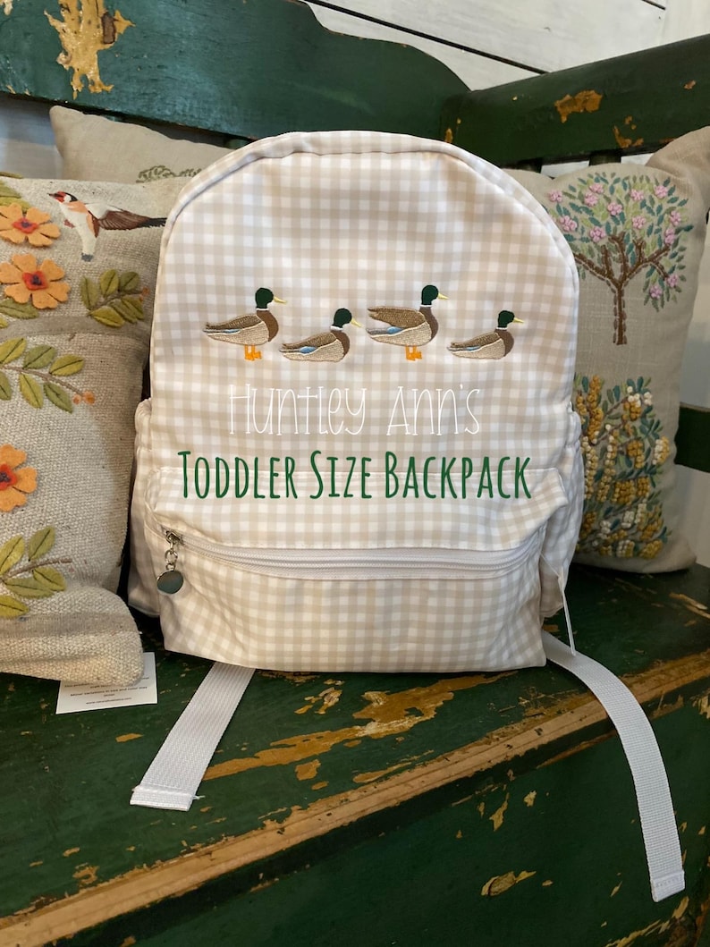 May include: A beige and white gingham toddler-sized backpack with a zipper closure. The backpack has embroidered ducks and the text "Huntley Ann's Toddler Size Backpack".