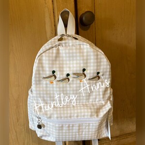 May include: A white and beige gingham backpack with a zippered pocket and embroidered ducks and the name "Huntley Ann's".