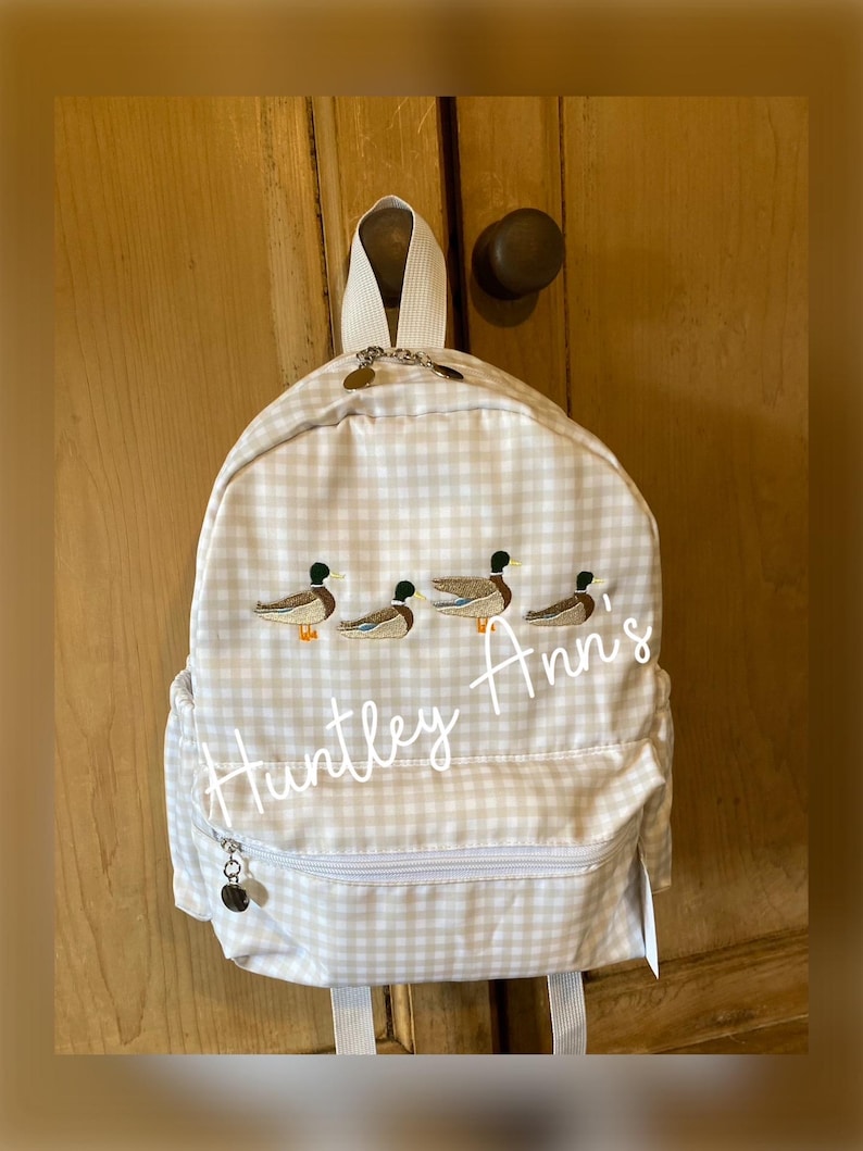 May include: A white and beige gingham backpack with a zippered pocket and embroidered ducks and the name "Huntley Ann's".