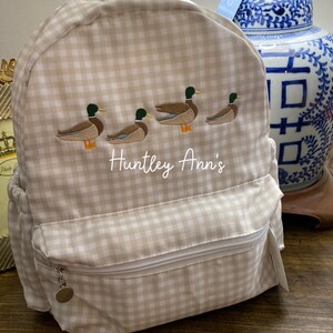 May include: A beige and white gingham backpack with a zipper closure and a small pocket on the front. The backpack has an embroidered design of four ducks in a row and the text "Huntley Ann's".