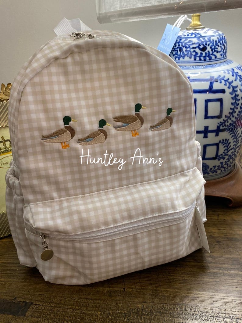 May include: A beige and white gingham backpack with a zipper closure and a small pocket on the front. The backpack has an embroidered design of four ducks in a row and the text "Huntley Ann's".