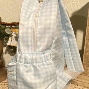 Puede incluir: Una mochila de cuadros vichy azul claro y blanco con cremallera blanca y tirador plateado en forma de coraz&oacute;n. La mochila tiene un bolsillo delantero y correas blancas. La bolsa es de material brillante.