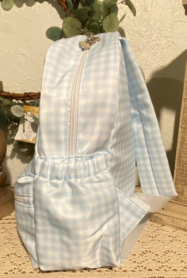 Puede incluir: Una mochila de cuadros vichy azul claro y blanco con cremallera blanca y tirador plateado en forma de coraz&oacute;n. La mochila tiene un bolsillo delantero y correas blancas. La bolsa es de material brillante.