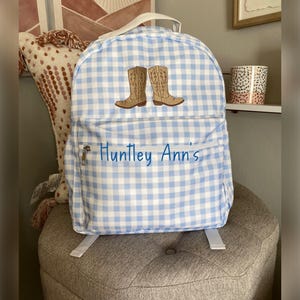 Puede incluir: Una mochila a cuadros azules y blancos con botas de vaquero bordadas. La mochila tiene un bolsillo frontal con cremallera con el nombre "Huntley Ann's" en escritura azul. Cuenta con un asa superior y correas ajustables.