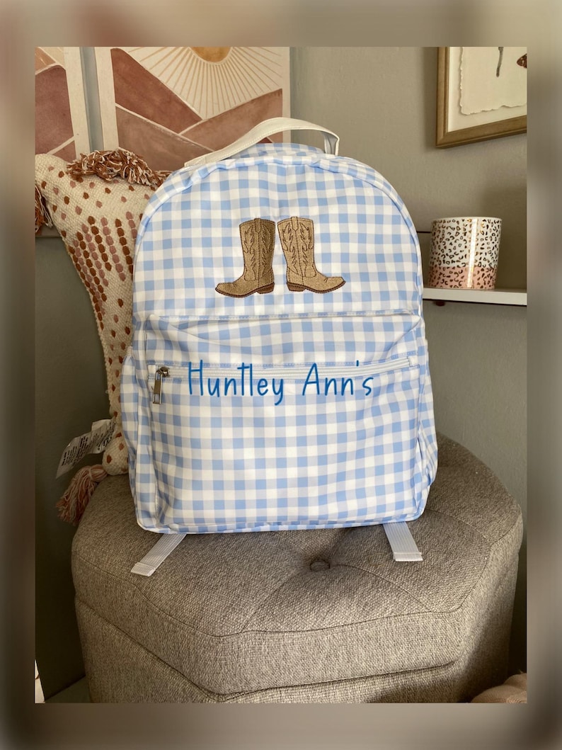 Puede incluir: Una mochila a cuadros azules y blancos con botas de vaquero bordadas. La mochila tiene un bolsillo frontal con cremallera con el nombre "Huntley Ann's" en escritura azul. Cuenta con un asa superior y correas ajustables.