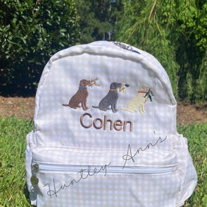 May include: A white and tan gingham backpack with embroidered dogs carrying sticks. The name "Cohen" is embroidered in brown thread above the front pocket. The name "Huntley Ann's" is written in cursive on the front.