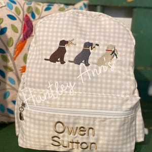 May include: A beige and white gingham backpack with embroidered images of three dogs carrying twigs. The backpack has a zippered front pocket and the name "Owen Sutton" embroidered in gold thread. The name "Huntley Ann's" is also embroidered.