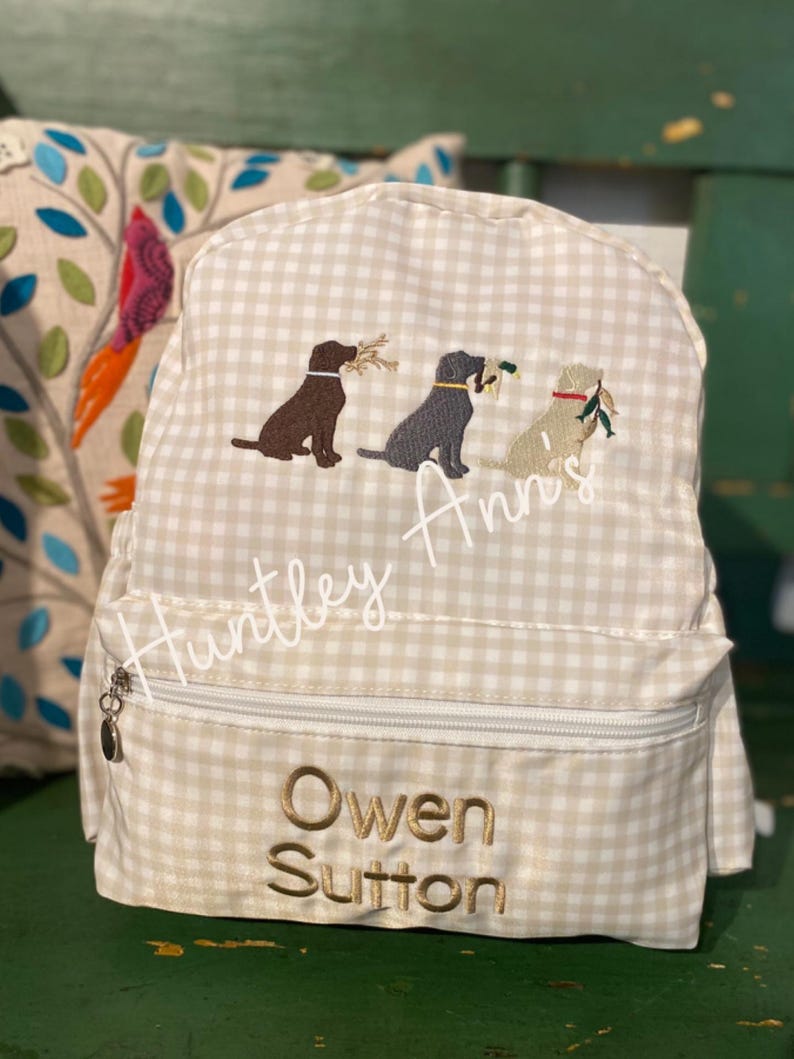 May include: A beige and white gingham backpack with embroidered images of three dogs carrying twigs. The backpack has a zippered front pocket and the name "Owen Sutton" embroidered in gold thread. The name "Huntley Ann's" is also embroidered.
