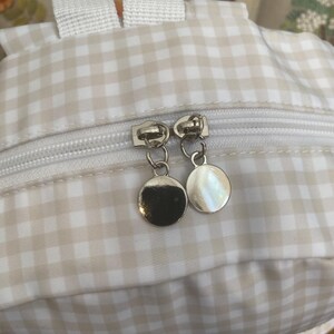 May include: Close-up of a small backpack with a beige and white gingham pattern. The backpack has a white zipper and white straps. The zipper pulls are silver, circular charms.