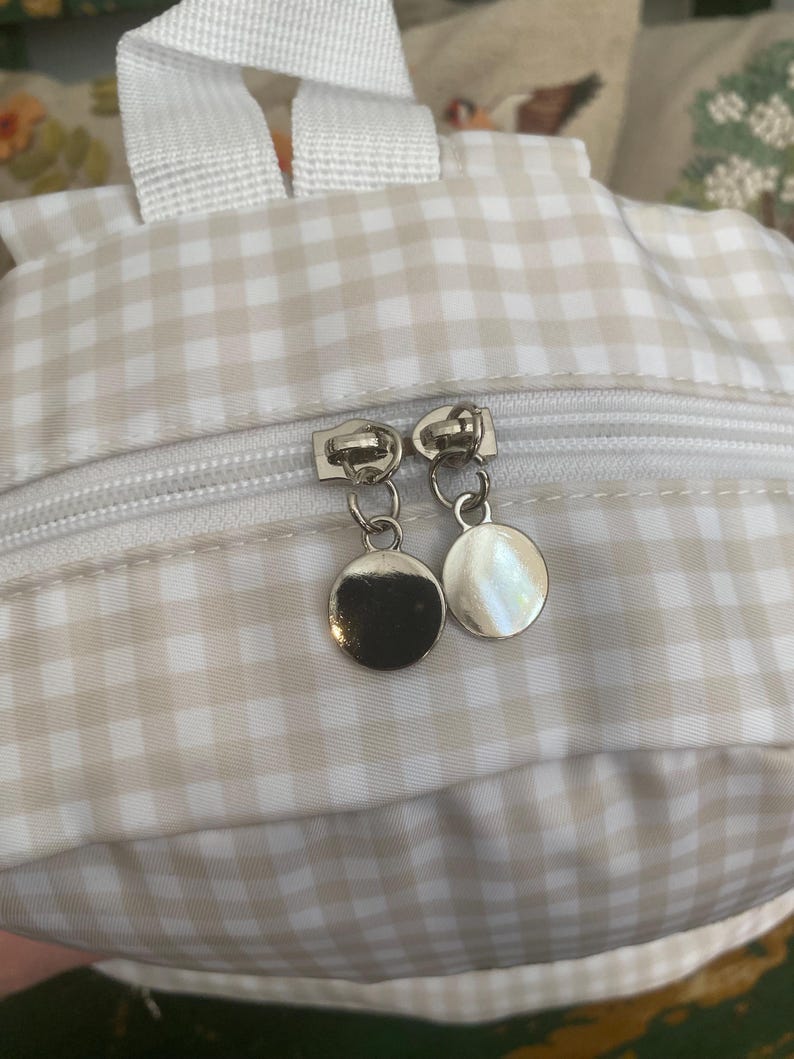 May include: Close-up of a small backpack with a beige and white gingham pattern. The backpack has a white zipper and white straps. The zipper pulls are silver, circular charms.