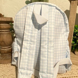 Puede incluir: Una mochila peque&ntilde;a con un patr&oacute;n de cuadros vichy azul claro y blanco, con correas blancas y un asa superior. Las correas ajustables para los hombros son visibles. La parte trasera de la bolsa muestra los detalles de costura.
