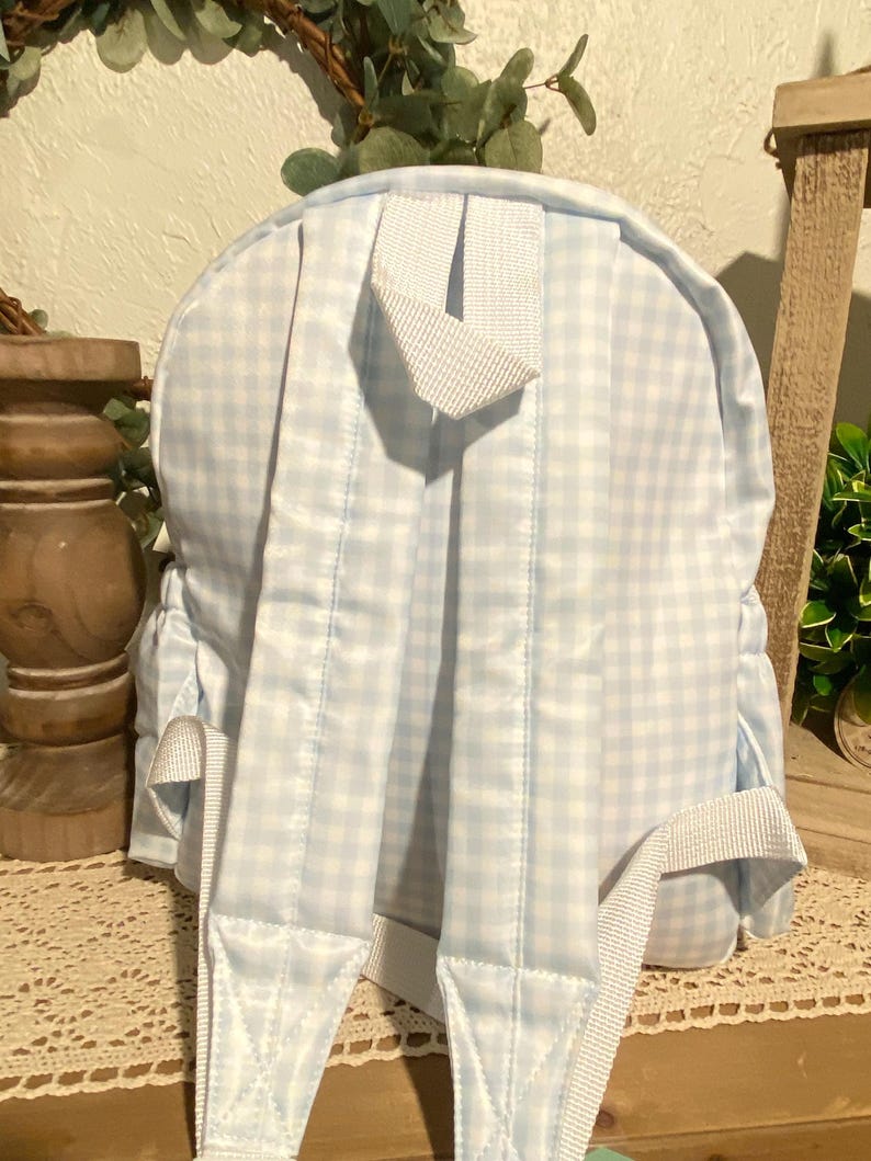 Puede incluir: Una mochila peque&ntilde;a con un patr&oacute;n de cuadros vichy azul claro y blanco, con correas blancas y un asa superior. Las correas ajustables para los hombros son visibles. La parte trasera de la bolsa muestra los detalles de costura.