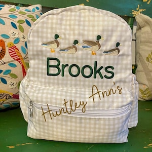 May include: A white and beige gingham fabric backpack with a zippered pocket. The backpack has embroidered ducks and the names "Brooks" and "Huntley Ann's".