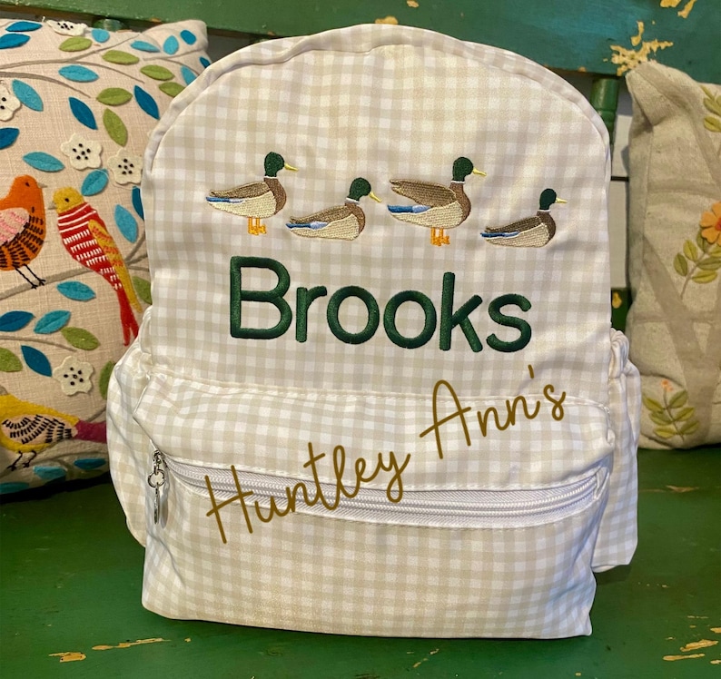 May include: A white and beige gingham fabric backpack with a zippered pocket. The backpack has embroidered ducks and the names "Brooks" and "Huntley Ann's".