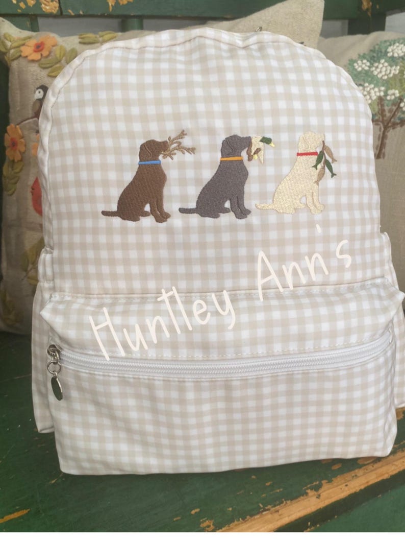 May include: A beige and white gingham backpack with three embroidered Labrador Retrievers carrying items in their mouths. The backpack has a front zippered pocket and the name "Huntley Ann's" embroidered on the front.