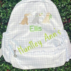 May include: Light blue and white gingham backpack with three embroidered dog illustrations, each holding a twig. The name "Ellis" is embroidered in green, with "Huntley Ann's" in a cursive font. Features a zippered front pocket.