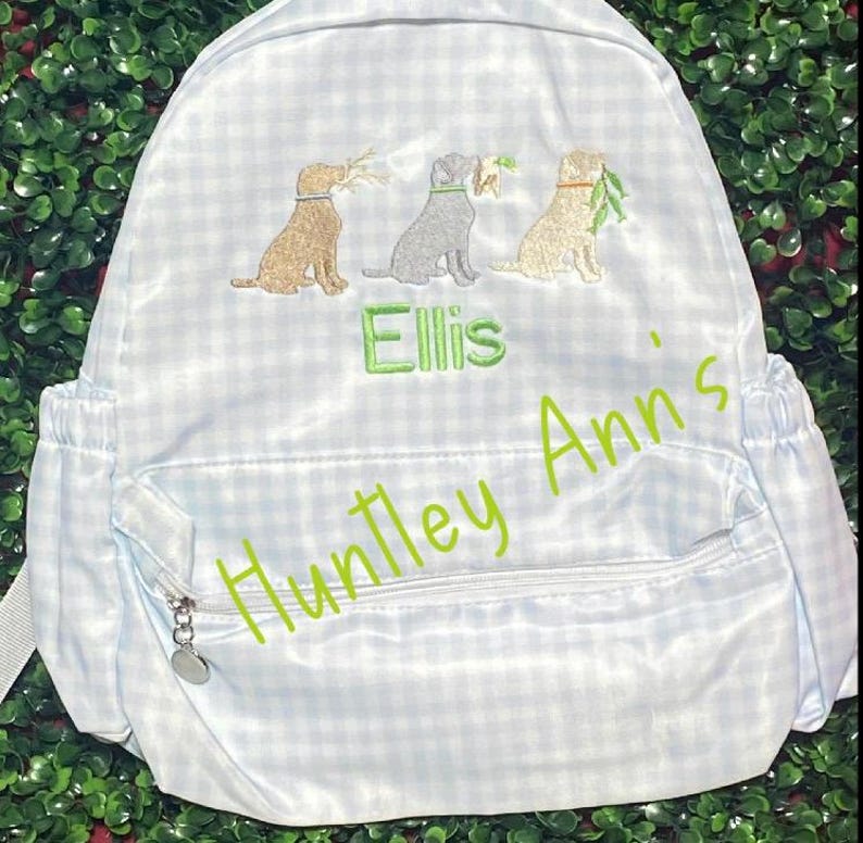 May include: Light blue and white gingham backpack with three embroidered dog illustrations, each holding a twig. The name "Ellis" is embroidered in green, with "Huntley Ann's" in a cursive font. Features a zippered front pocket.