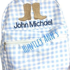Puede incluir: Una mochila a cuadros azul claro y blanco con un bolsillo con cremallera y un asa superior. La mochila presenta un dise&ntilde;o bordado de dos botas de vaquero marrones y el nombre "John Michael" en azul marino. Tambi&eacute;n es visible el texto "HUNTLEY ANN'S".