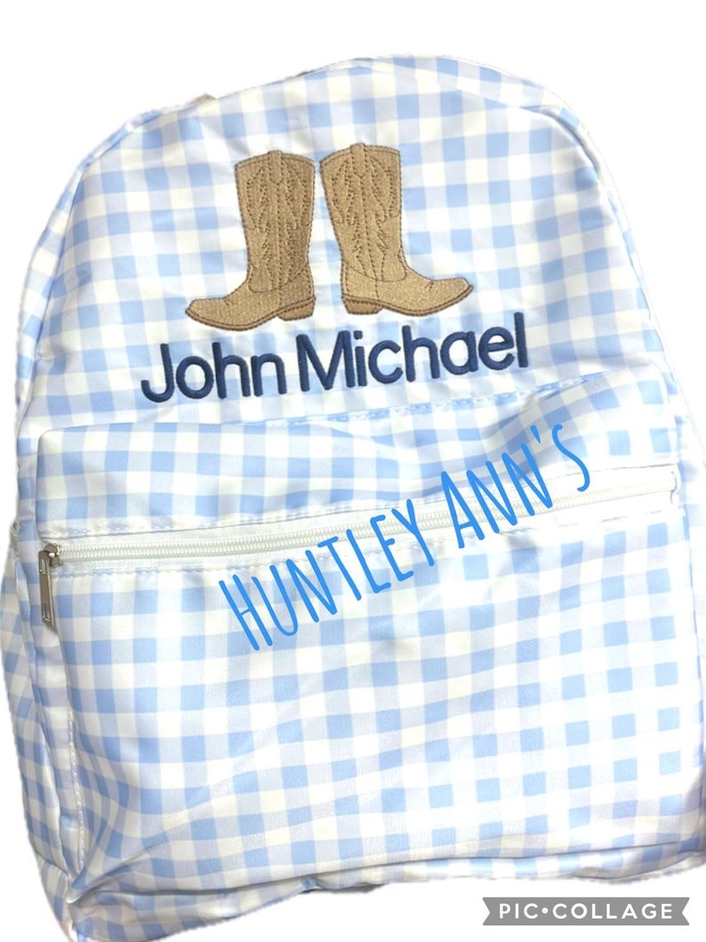 Puede incluir: Una mochila a cuadros azul claro y blanco con un bolsillo con cremallera y un asa superior. La mochila presenta un dise&ntilde;o bordado de dos botas de vaquero marrones y el nombre "John Michael" en azul marino. Tambi&eacute;n es visible el texto "HUNTLEY ANN'S".