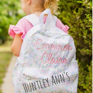 May include: A white floral backpack with the names "Emerson Claire" embroidered in pink. The text "HUNTLEY ANN'S" is printed on the front. The backpack has a zipper closure and white straps. The background is a blurred green.