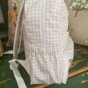 May include: A beige and white gingham backpack with a white zipper and straps. The backpack has a small side pocket with a zipper and a front pocket. The background includes decorative pillows.