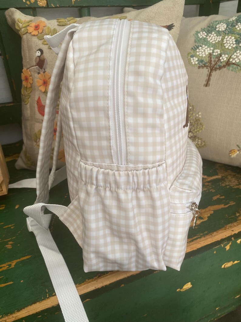May include: A beige and white gingham backpack with a white zipper and straps. The backpack has a small side pocket with a zipper and a front pocket. The background includes decorative pillows.