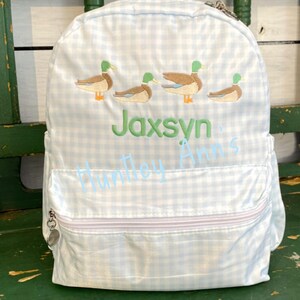 May include: A blue and white gingham backpack with a zipper closure. The backpack has embroidered ducks and the name "Jaxsyn" in green.