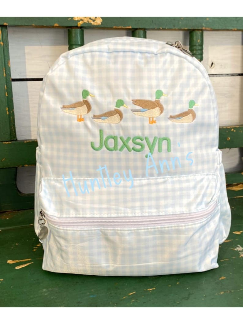 May include: A blue and white gingham backpack with a zipper closure. The backpack has embroidered ducks and the name "Jaxsyn" in green.