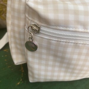 May include: Close-up of a beige and white gingham pattern backpack with a silver zipper and a round charm. The bag has a pocket with a zipper closure and a white strap. The fabric is a light beige and white checkered pattern.