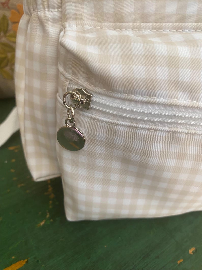 May include: Close-up of a beige and white gingham pattern backpack with a silver zipper and a round charm. The bag has a pocket with a zipper closure and a white strap. The fabric is a light beige and white checkered pattern.