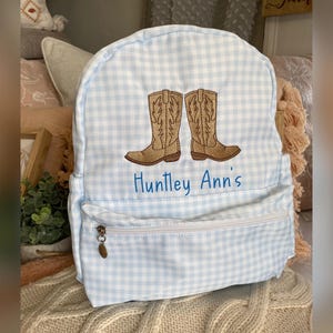 Puede incluir: Una mochila a cuadros vichy azul claro y blanco con un dise&ntilde;o bordado de dos botas de vaquero marrones. El texto "Huntley Ann's" est&aacute; bordado en azul debajo. La mochila tiene un bolsillo frontal con cremallera.