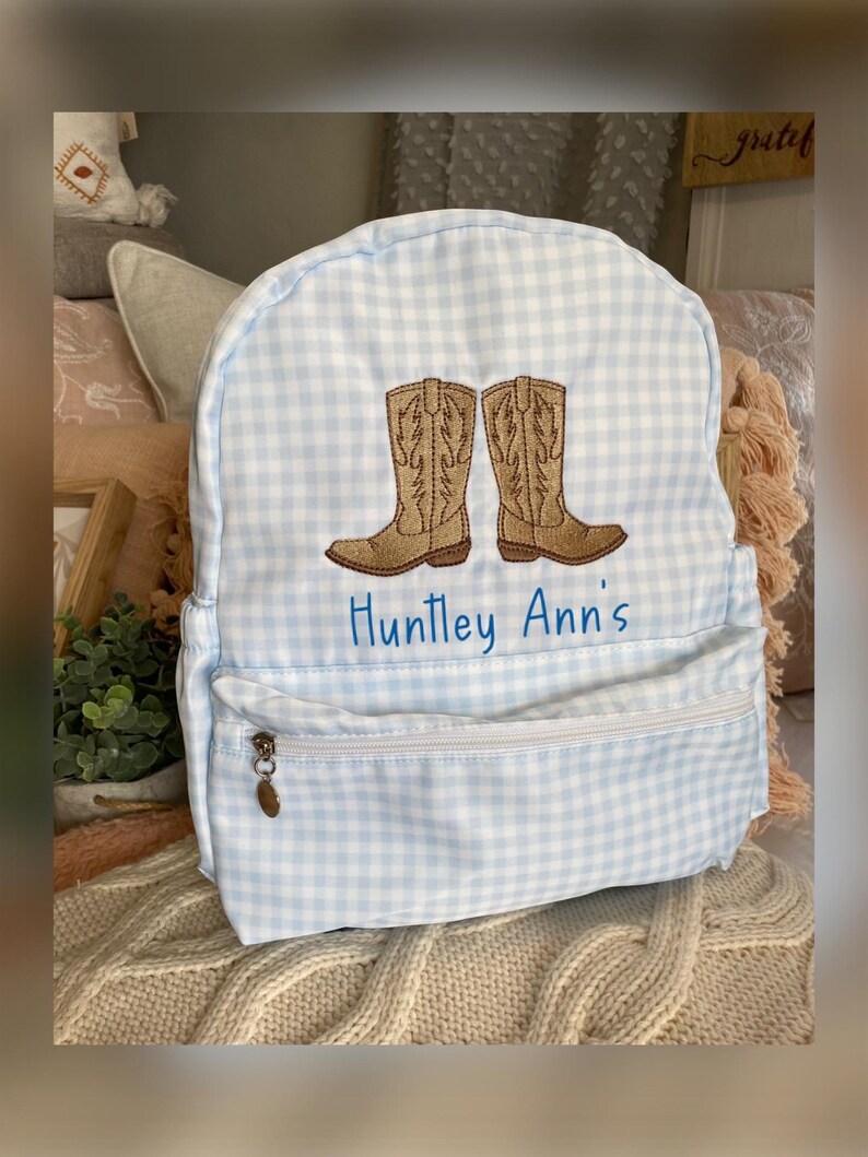 Puede incluir: Una mochila a cuadros vichy azul claro y blanco con un dise&ntilde;o bordado de dos botas de vaquero marrones. El texto "Huntley Ann's" est&aacute; bordado en azul debajo. La mochila tiene un bolsillo frontal con cremallera.