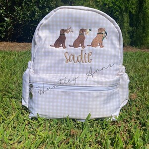 May include: A beige and white gingham backpack with three embroidered brown Labrador retrievers carrying items. The name "Sadie" is embroidered in gold thread above the dogs. The text "Huntley Ann's" is written in cursive on the front.
