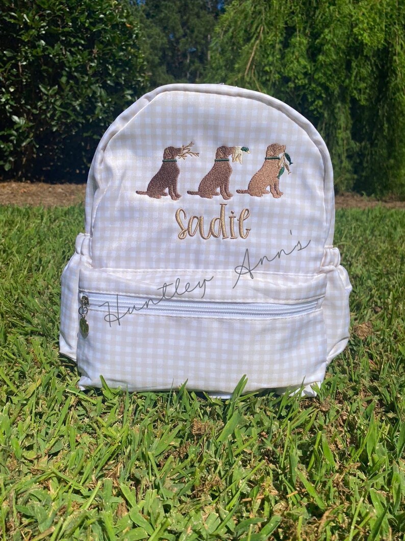 May include: A beige and white gingham backpack with three embroidered brown Labrador retrievers carrying items. The name "Sadie" is embroidered in gold thread above the dogs. The text "Huntley Ann's" is written in cursive on the front.