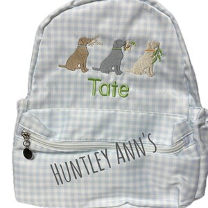 May include: A light blue and white gingham backpack with three embroidered dogs carrying foliage. The backpack has a front zippered pocket and the text "Huntley Ann's" and "Tate".