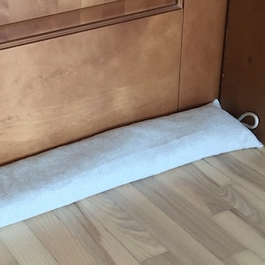 Può includere: Un paraspifferi in tessuto grigio chiaro, di forma rettangolare, posto alla base di una porta in legno. Il paraspifferi è progettato per bloccare le correnti d'aria e risparmiare energia. La porta è marrone chiaro.