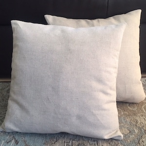May include: Two square throw pillows featuring a plain, light beige, woven fabric cover. The pillows have a simple, clean design with a visible texture in the weave, presented in a stacked arrangement with one pillow positioned slightly behind the other.