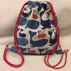Könnte beinhalten: Ein Kordelzugrucksack aus beigem Stoff mit einem sich wiederholenden Muster aus Walen in Marineblau, Hellblau und Orange. Der Beutel wird oben mit leuchtend roten, geflochtenen Kordelzügen verschlossen, die an den unteren Ecken befestigt sind.