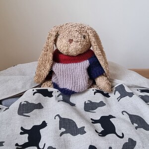 May include: A plush tan rabbit toy with long floppy ears wearing a knitted jumper with horizontal bands of red, light purple, and dark blue. The rabbit is positioned behind a light beige fabric featuring a repeating pattern of black cat silhouettes.