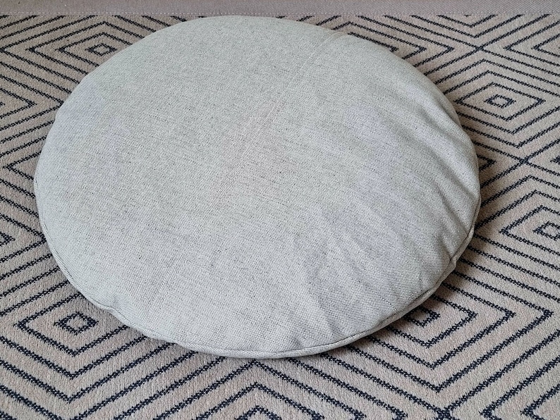 Op de afbeelding: Een rond, lichtgrijs, stoffen kussen met een gladde textuur. Het kussen ligt op een tapijt met een patroon.