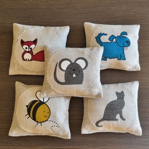 May include: Five square beige linen sachets featuring circular patches with screen-printed animal illustrations. The designs include a red fox, a blue dog, a grey mouse, a yellow and black bee, and a grey cat, each centred on the fabric surface.