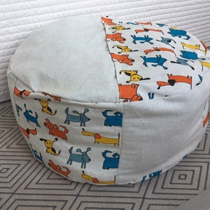 Peut inclure: Un pouf de sol rond composé de panneaux alternés en tissu beige clair uni et en tissu à motifs représentant des chiens de dessin animé orange, bleus et jaunes. Le pouf présente une forme cylindrique structurée avec des coutures visibles.