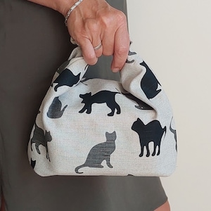 Op de afbeelding: Een kleine handtas van beige stof met een herhalend patroon van zwarte katten-silhouetten in verschillende houdingen. De tas heeft een zachte, ongestructureerde vorm met geïntegreerde handvatten gevormd door de geplooide bovenstof.