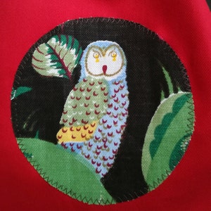 May include: A round, black and green fabric patch with a white owl with yellow eyes. The owl is surrounded by green leaves and a black background.