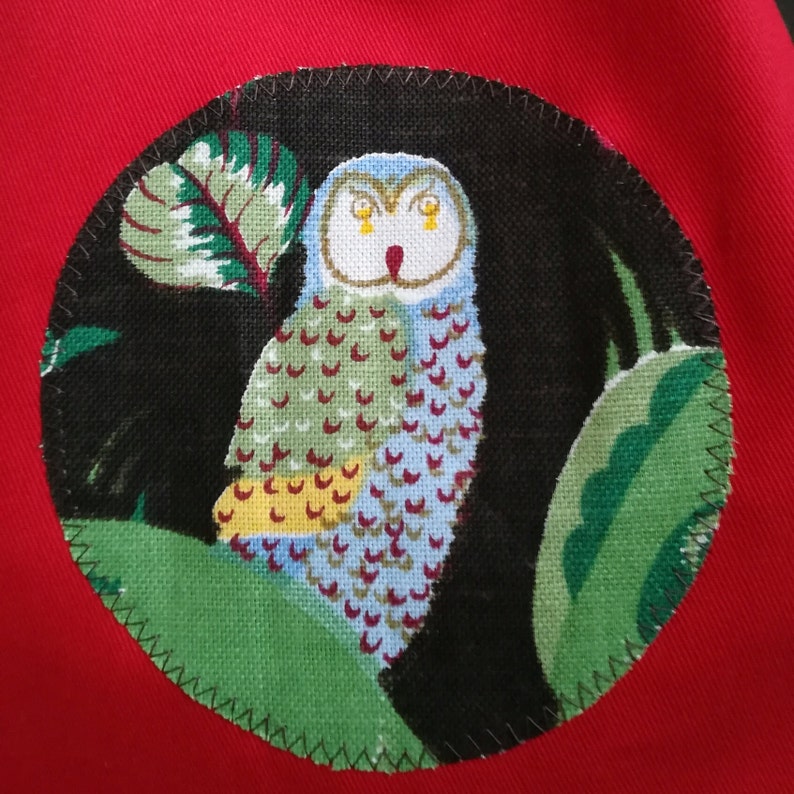 May include: A round, black and green fabric patch with a white owl with yellow eyes. The owl is surrounded by green leaves and a black background.