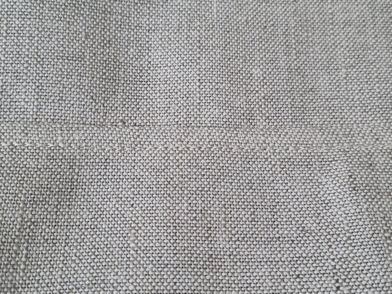 Op de afbeelding: Close-up van een lichtgrijze geweven stof met een subtiele textuur. De stof heeft een gladde, gelijkmatige weving en een licht ruw gevoel.